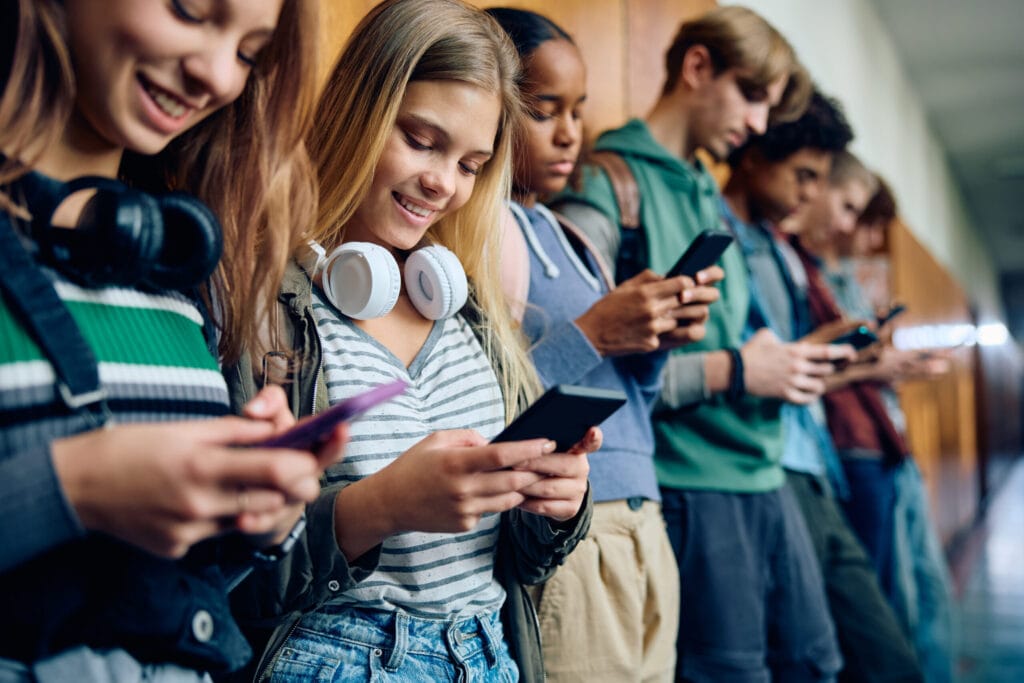 A group of diverse teenage classmates smiling and using their smartphones in a high school hallway, capturing social media use on campus before California's statewide restrictions went into effect.