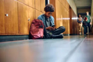 A Black teenage high school student texting on his smartphone in a school hallway, illustrating cell phone distraction before California's AB 3216 phone-free schools ban takes effect.