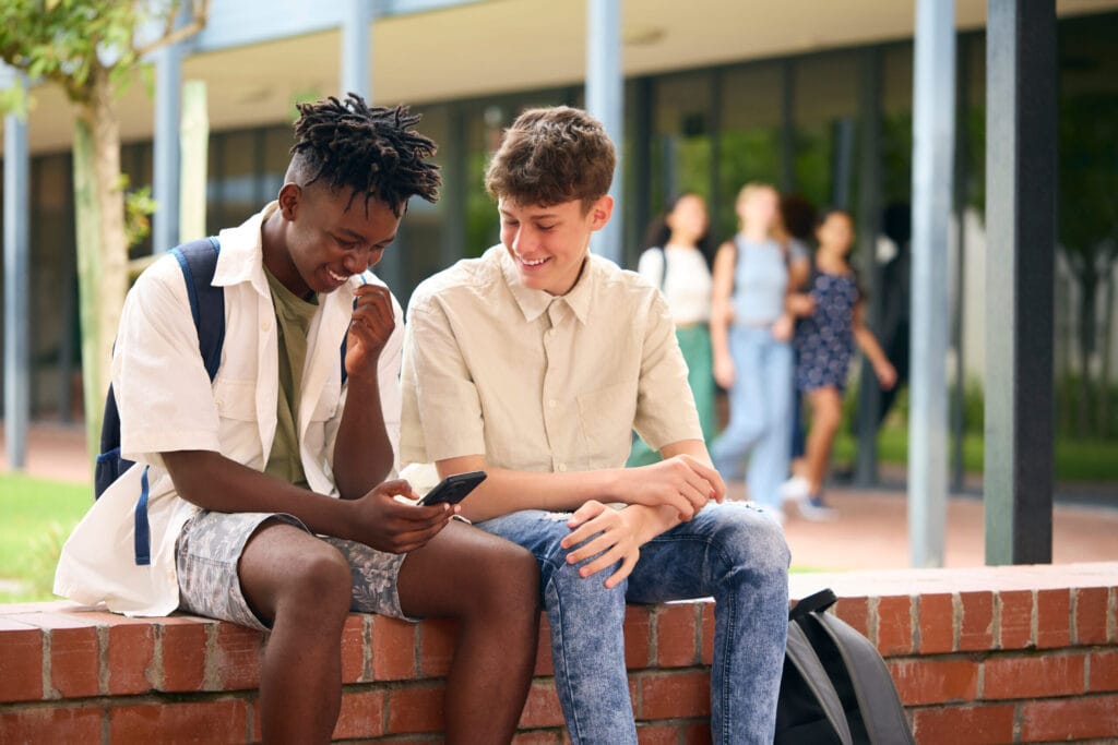 Two male high school students looking at a smartphone screen together in an outdoor courtyard, highlighting the peer-to-peer digital distraction targeted by the California Phone-Free Schools Act.